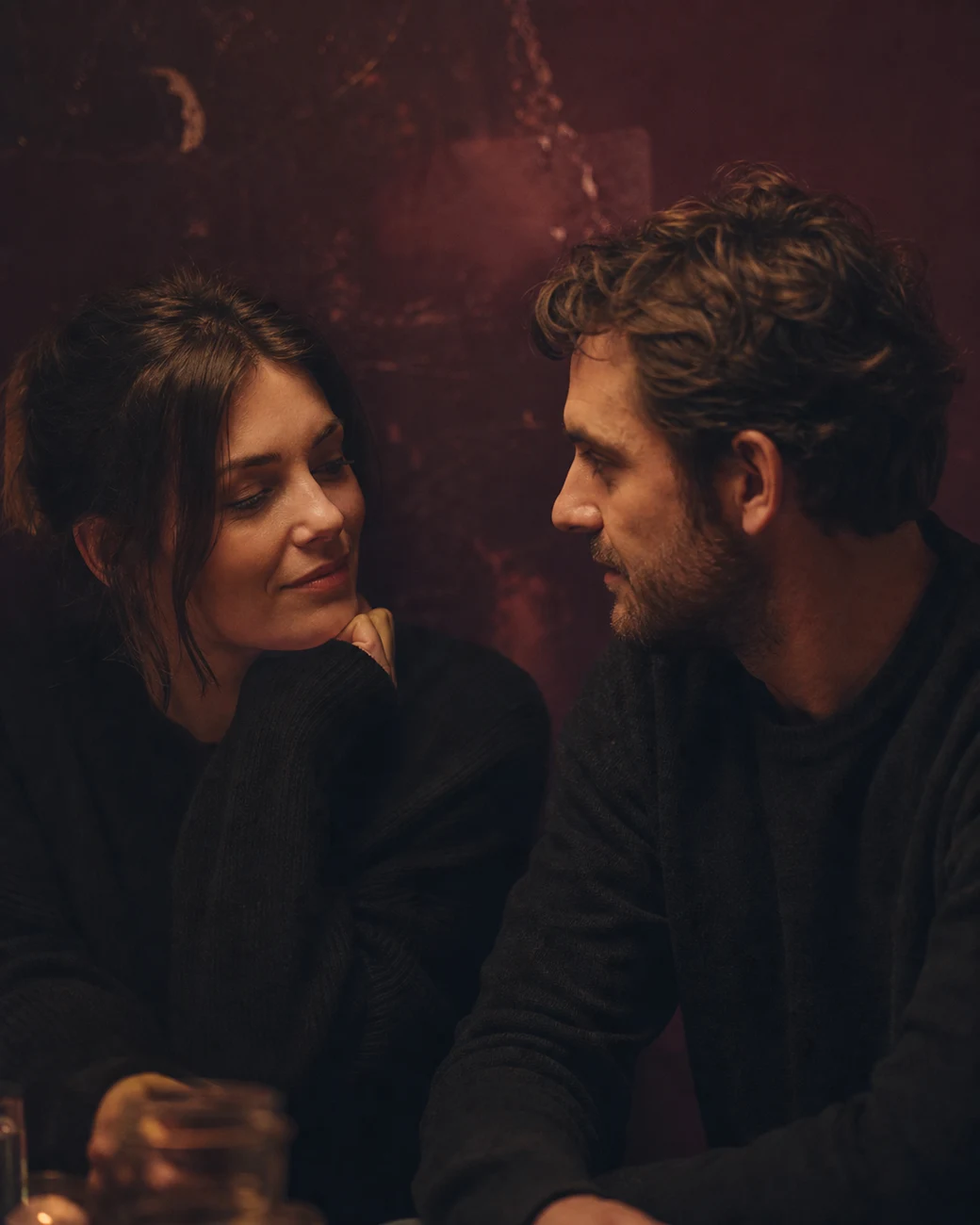 Two people sitting close in warm candlelight, turned toward each other mid-conversation.